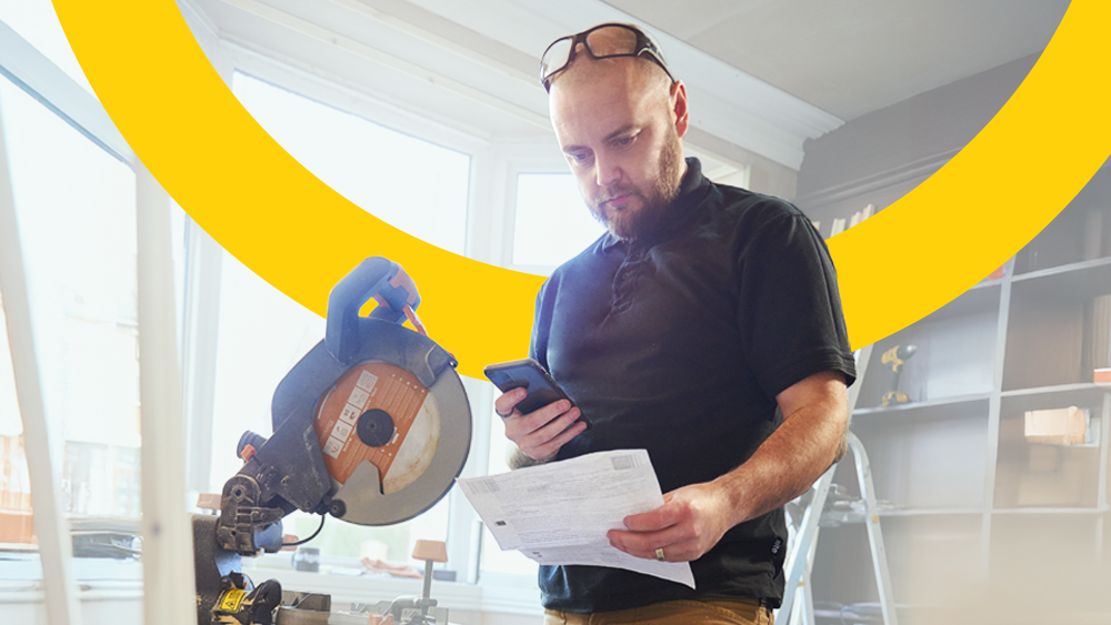 Joiner looking at their mobile phone with paper documents for Making Tax Digital